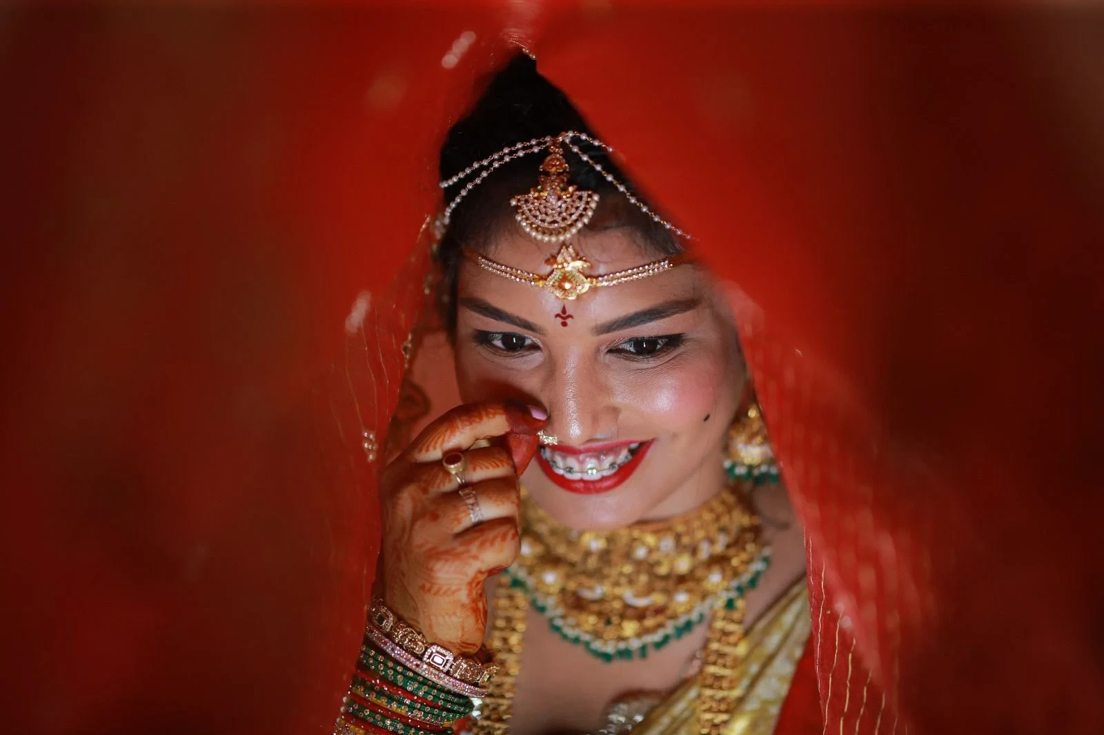 Bride smiling beneath a red veil, wearing traditional jewellery and bridal makeup by Bindu